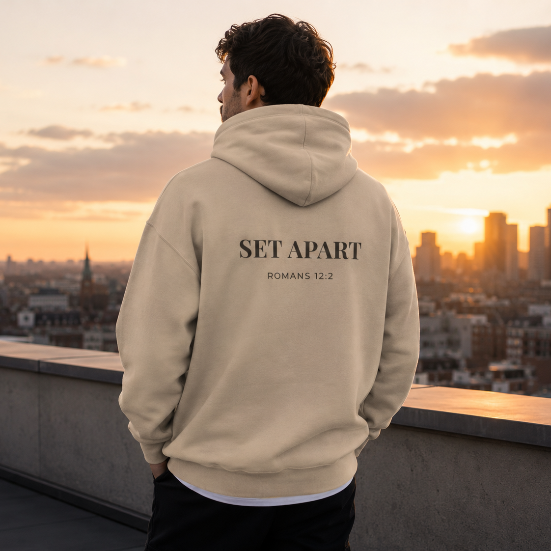 Person wearing a beige Cornerstone wear hoodie with 'SET APART' text, standing on a rooftop with a cityscape and sunset in the background.