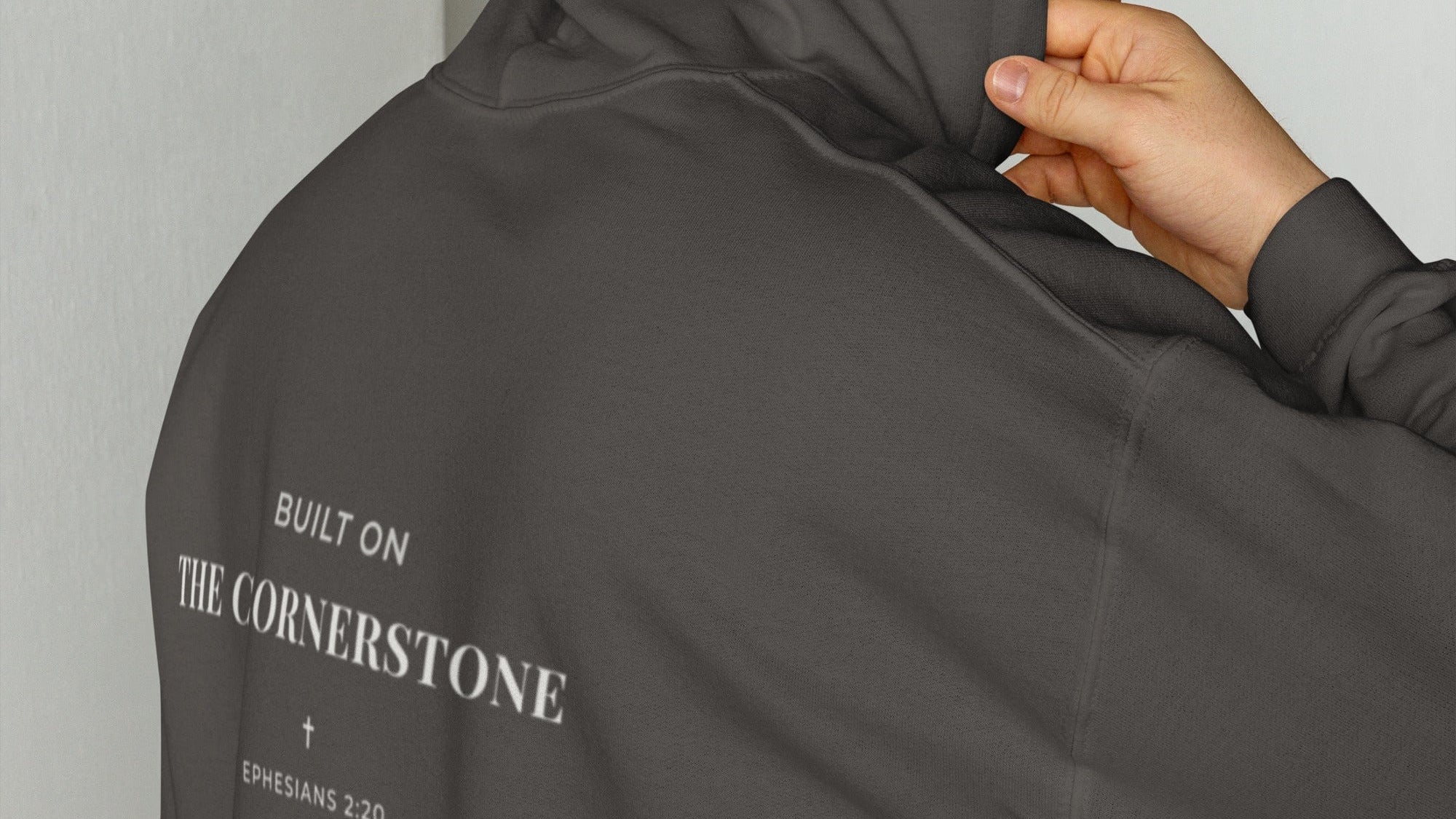Person wearing a Cornerstone Wear unisex dark grey hoodie with text, standing against a plain background