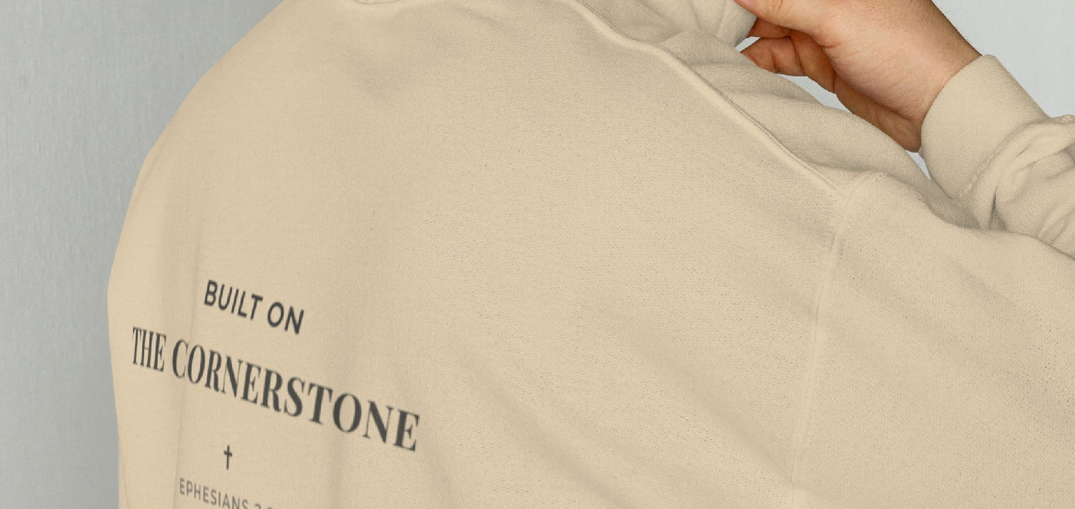 Person wearing a Cornerstone Wear unisex sand hoodie with text, standing against a plain background